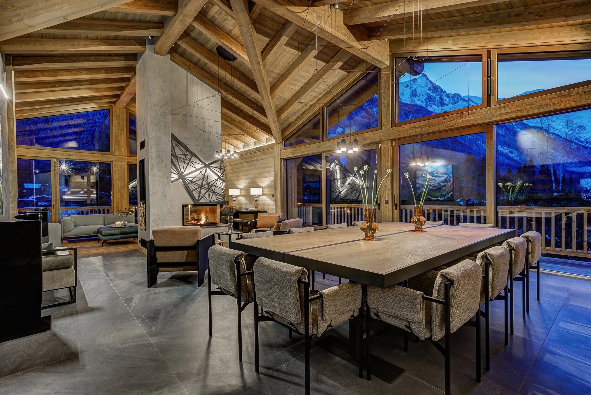 Luxury dining area inside Chalet Mélèzes set for a dinner with a large oak table, chairs, warm lighting, and floor-to-ceiling windows showing snow-covered mountains under a deep blue twilight sky